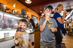 Two children enjoying ice cream cones inside a colorful ice cream shop in Spooner, Wisconsin, with a woman in the background holding a cone, highlighting local culinary delights.