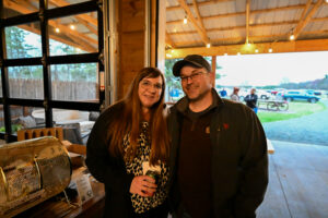 Couple smiling and posing for a photo at a community event, with a rustic venue and outdoor gathering visible in the background, reflecting the social atmosphere of the Spooner Area Chamber of Commerce Food & Wine Tasting Benefit.