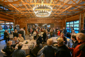 Crowd of attendees enjoying the Food & Wine Tasting Benefit at a rustic venue with a chandelier, mingling around tables and engaging in conversation.
