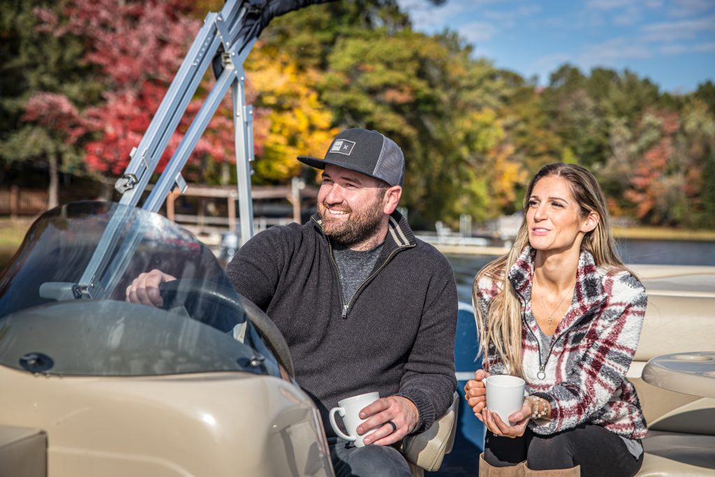 Couple enjoying a pontoon ride on Spooner Lake, surrounded by vibrant fall foliage, with the man steering the boat and the woman holding a coffee cup.