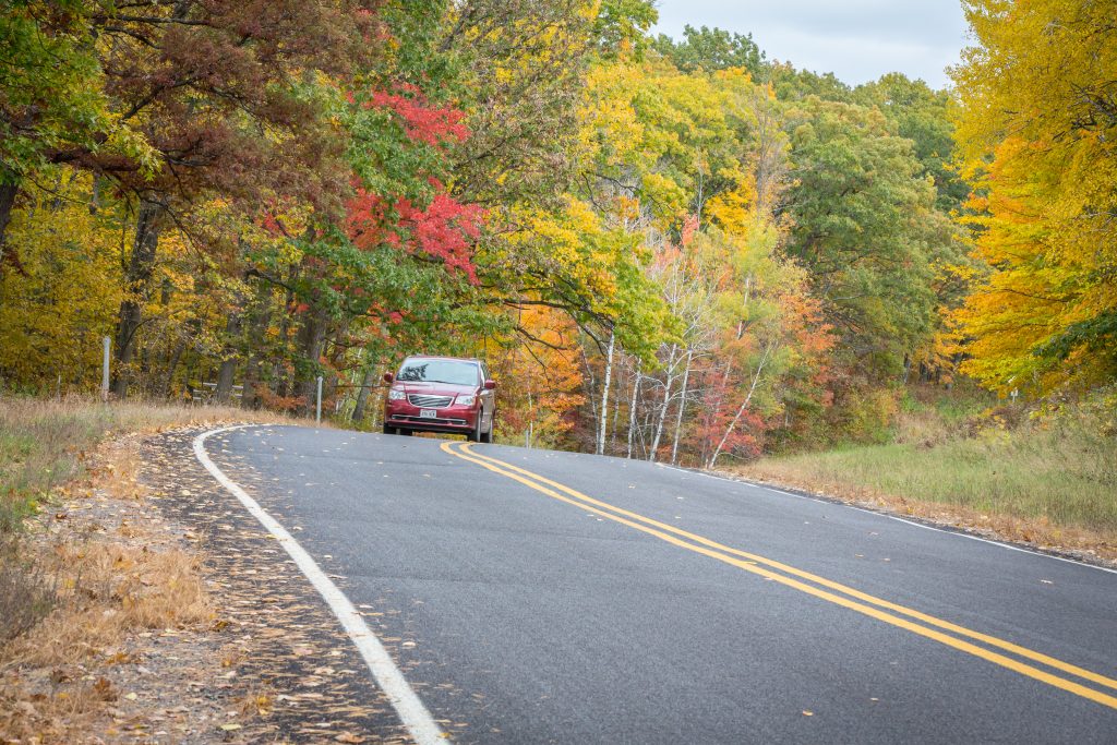 Curving road in Spooner, Wisconsin, lined with vibrant autumn foliage in shades of red, orange, and yellow, featuring a car navigating the scenic landscape.